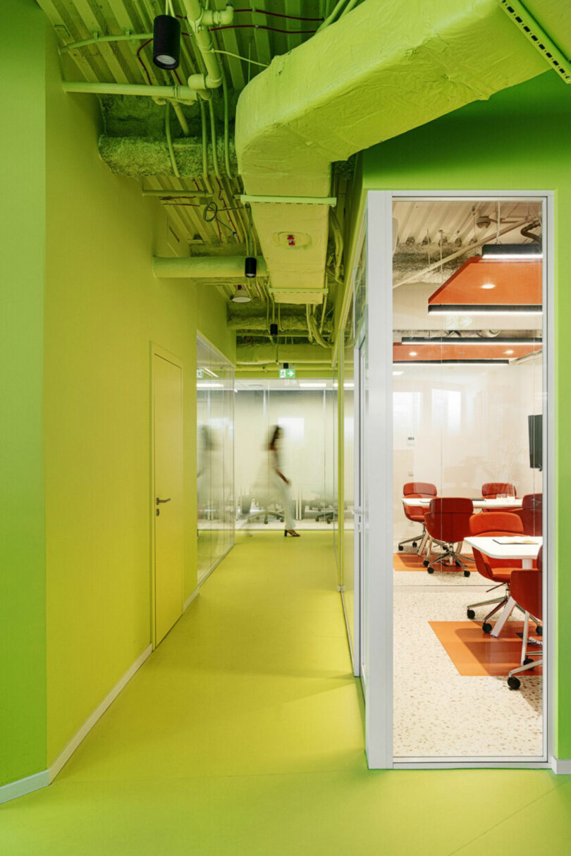 A brightly lit, modern office hallway with green walls and ceiling. Glass doors on the right open into a conference room with red chairs. Workers are visible through the frosted glass.