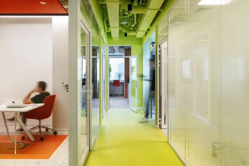 Modern office hallway with glass partition walls and a bright yellow floor, leading to an open room. A person is seated on the left, using a laptop at a desk.