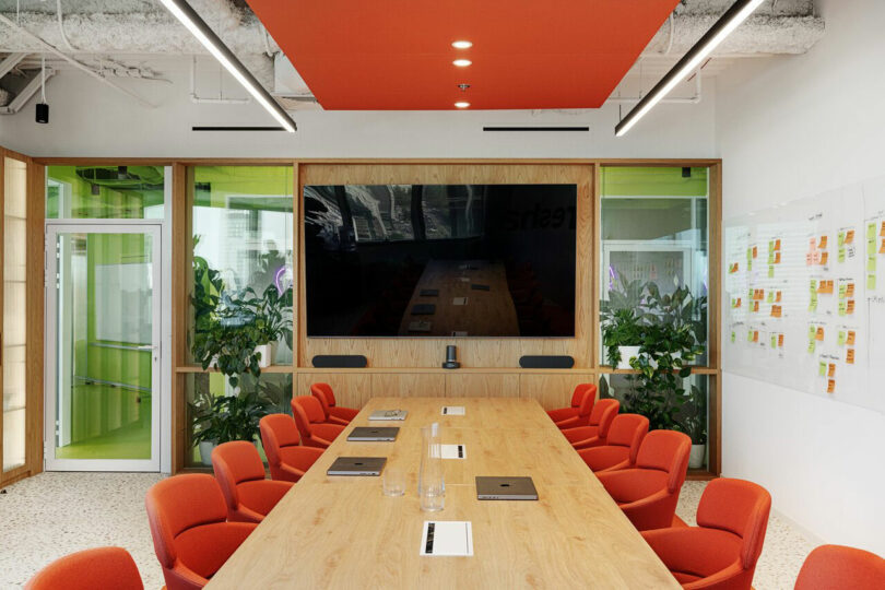 A modern conference room with a long wooden table, red chairs, large TV screen on the wall, and glass writing boards. The room has ceiling lights and green plants in the corners.