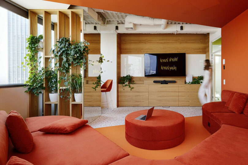 Modern lounge with red seating, a round ottoman, wooden shelves with plants, a wall-mounted TV, and a person walking by.