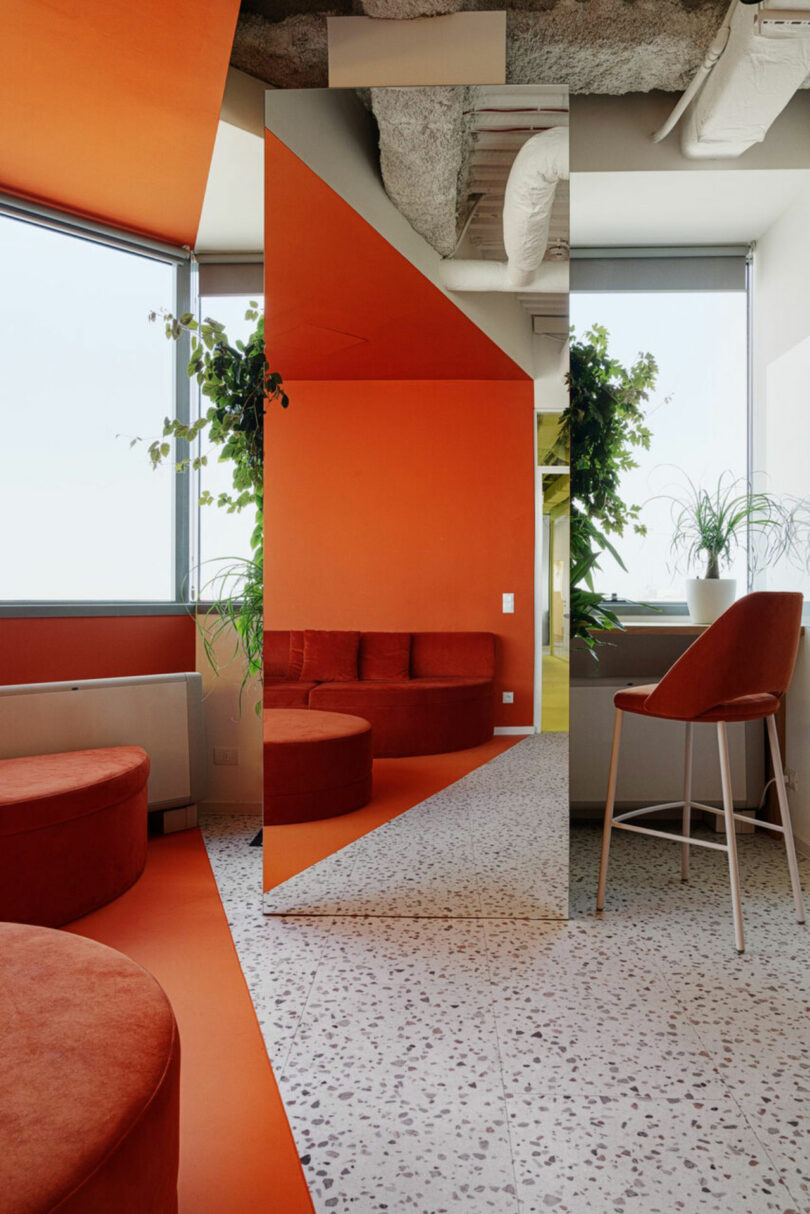 A modern room with orange walls, round seats, a tall mirror, and terrazzo flooring. Green plants by the windows and a high chair complete the setting.