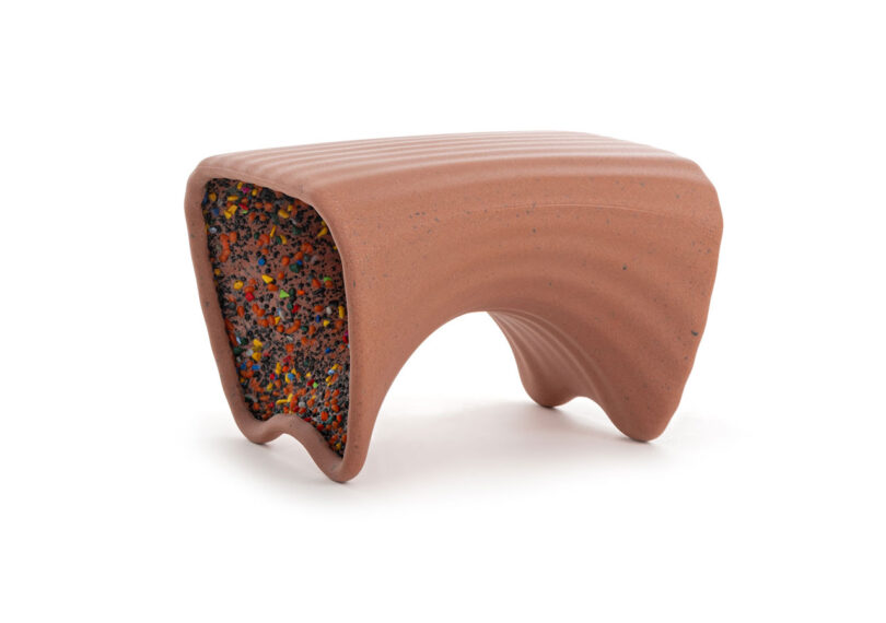 A small, curved bench inspired by In-Side Thomas Heatherwick, with a ribbed, terracotta-like exterior and a colorful, speckled interior surface, set against a white background.