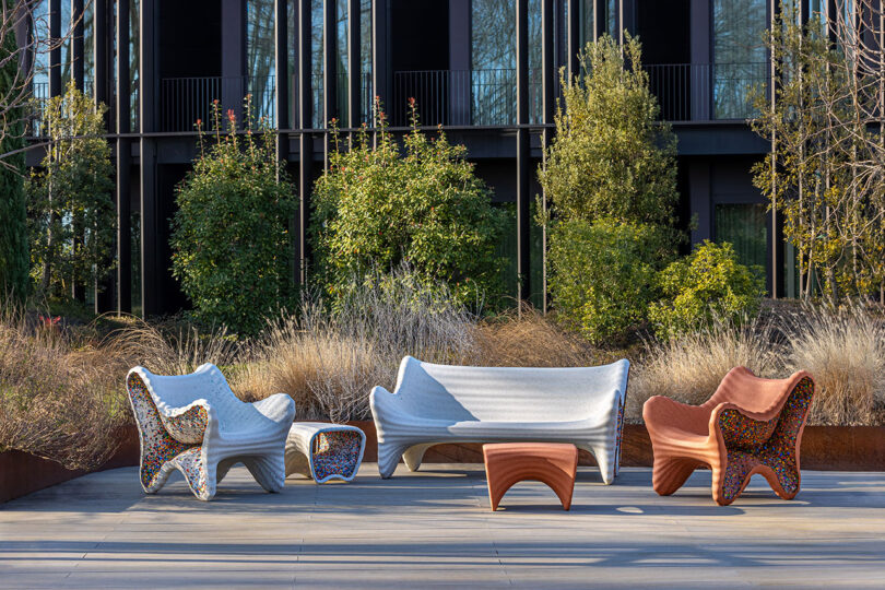 Outdoor seating area with uniquely designed chairs and a table on a patio, surrounded by greenery and a modern building in the background.