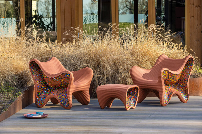 Two sculptural outdoor chairs and a matching table from the In-Side Thomas Heatherwick collection, all with wavy terracotta forms and colorful mosaic interiors, are placed on a patio near ornamental grasses.