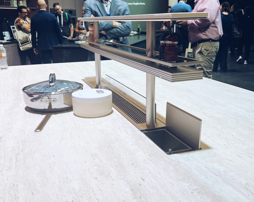 A sleek KBIS kitchen island features a built-in retractable shelf, a pot with lid, a white container, and a pull-out drawer. In the background, people are gathered around.