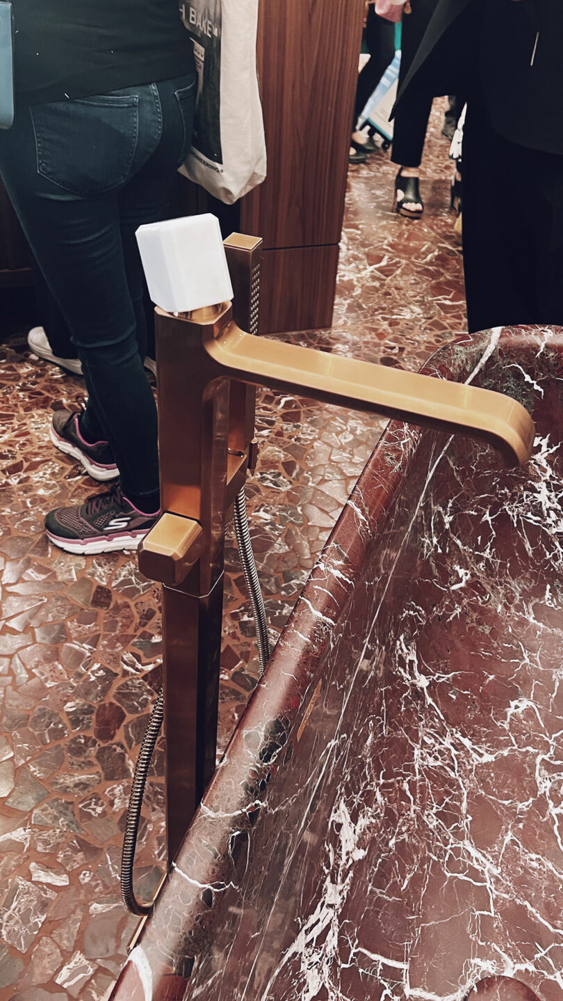 The bronze faucet with a white handle complements the red marble sink, all of which echoes the sophistication often showcased at KBIS. In the background, legs are visible on the tiled floor, completing this elegant scene.