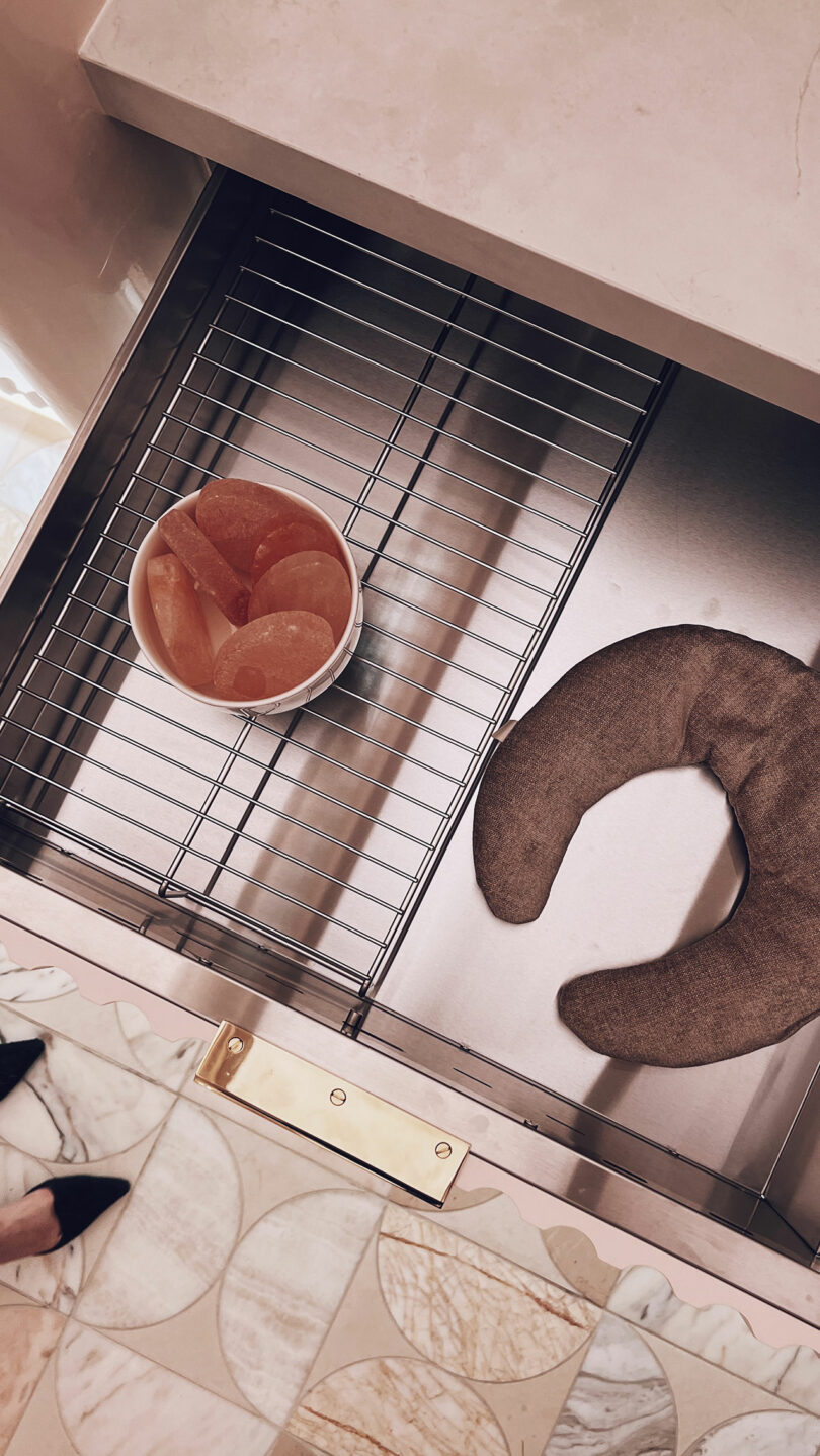 A bowl with orange slices sits on a metal rack in the kitchen sink, showcasing its freshness, reminiscent of a KBIS kitchen design. A brown crescent-shaped item is nearby, and part of the tiled floor is visible.