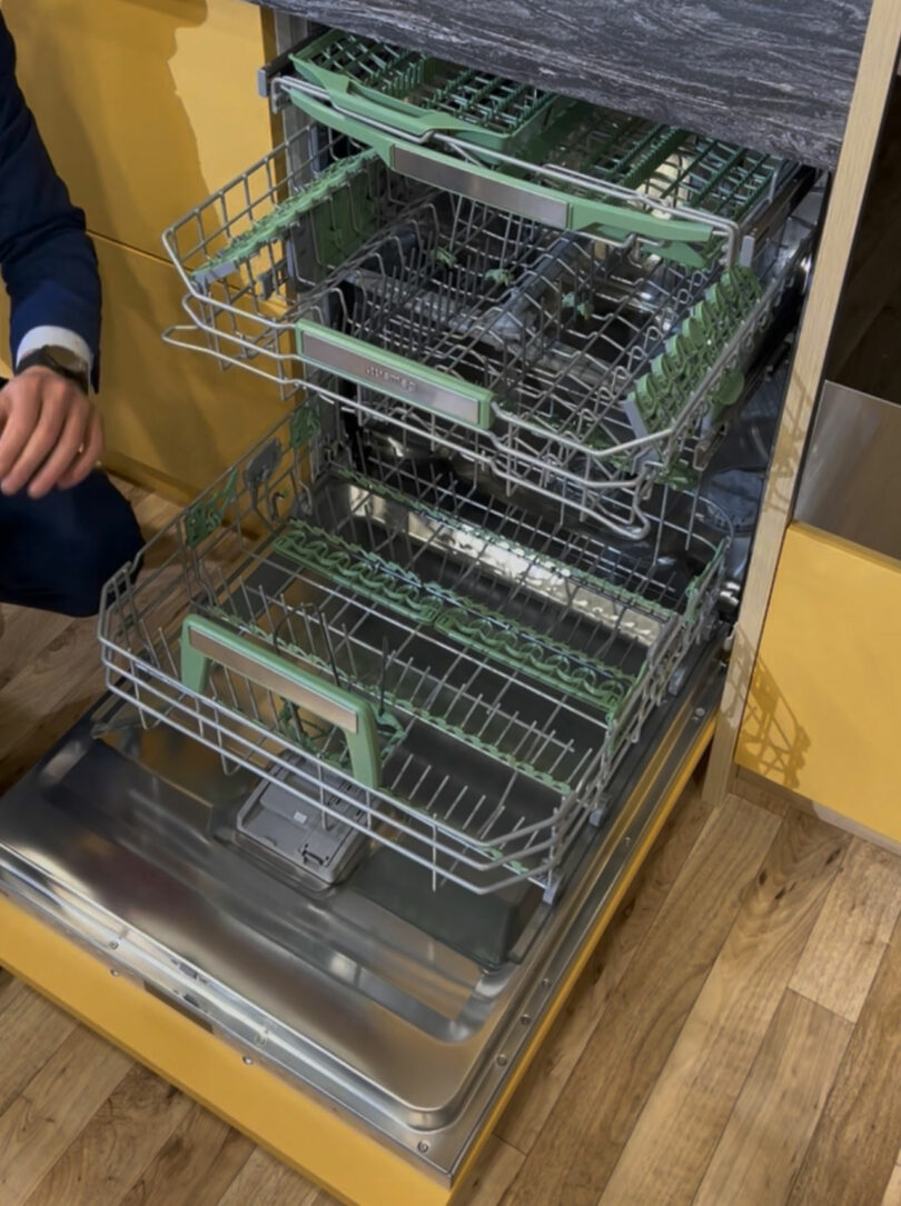 A person showcases a three-rack dishwasher with the door open, revealing the spacious interior racks and compartments, just like what you'd expect to see at a KBIS exhibit. The appliance is seamlessly integrated into a stylish wooden kitchen setup.