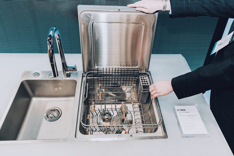 At KBIS, a person in a suit effortlessly demonstrates a compact, top-loading dishwasher seamlessly integrated into the countertop next to the sink.