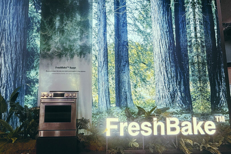 A stovetop and oven stand against a backdrop featuring tall trees and foliage, with the illuminated "FreshBake" text at the bottom, capturing the innovative spirit showcased at KBIS.