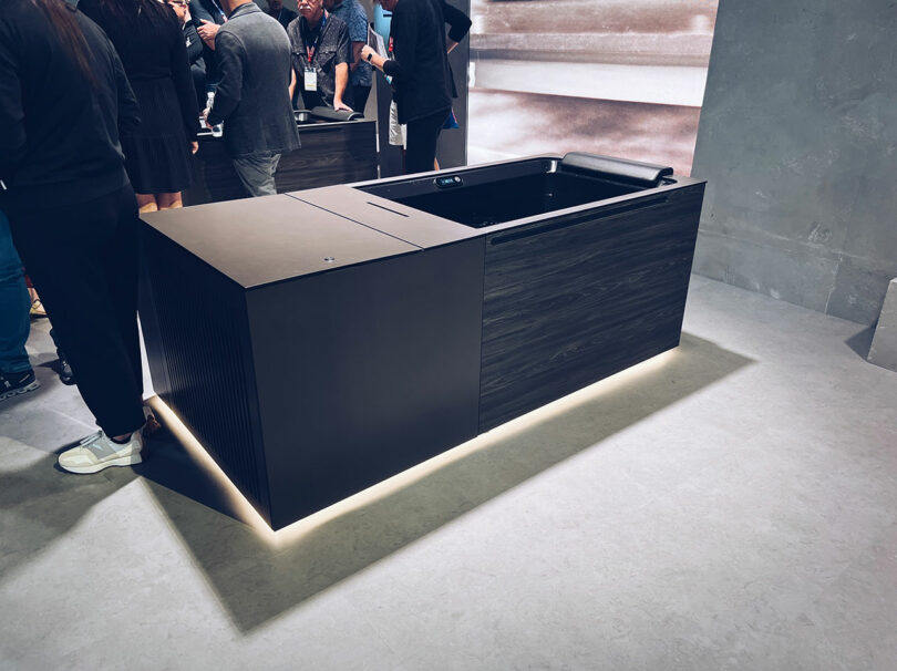 A modern, rectangular kitchen island with a sleek design and an attached sink becomes the centerpiece at KBIS. People are gathered around it, admiring its style and functionality.