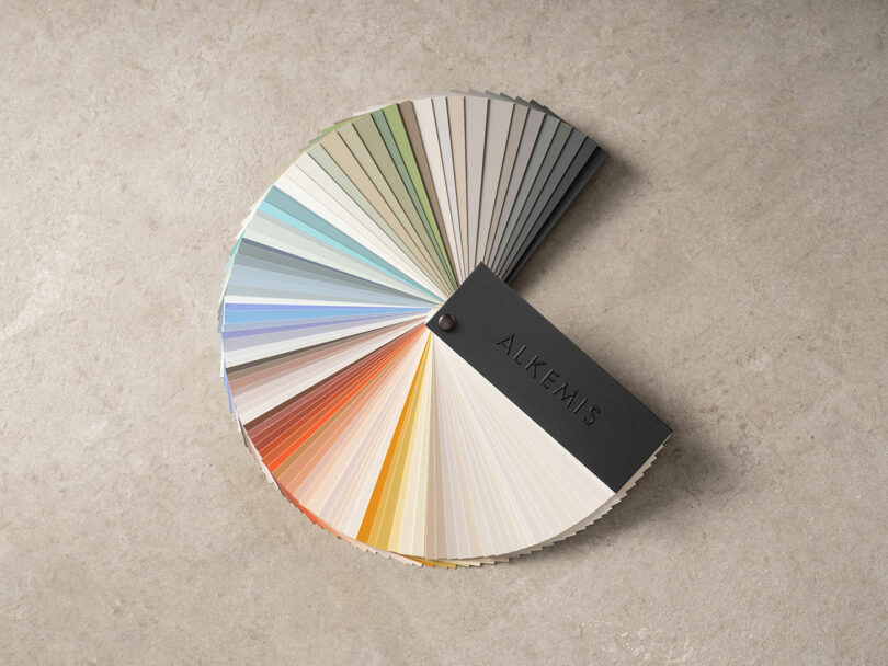 A spread of color swatches, curated for the discerning eye at KBIS, is gracefully arranged in a semicircle on a stone surface. Each hue whispers creativity beneath a sleek black cover labeled "ALKEMIS.