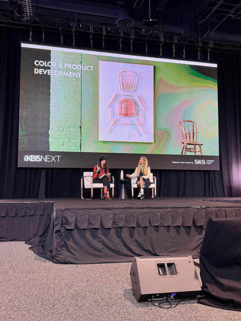 At a conference stage, two people engage in a discussion about color and product development at KBIS, with chair designs artfully displayed on the screen behind them.