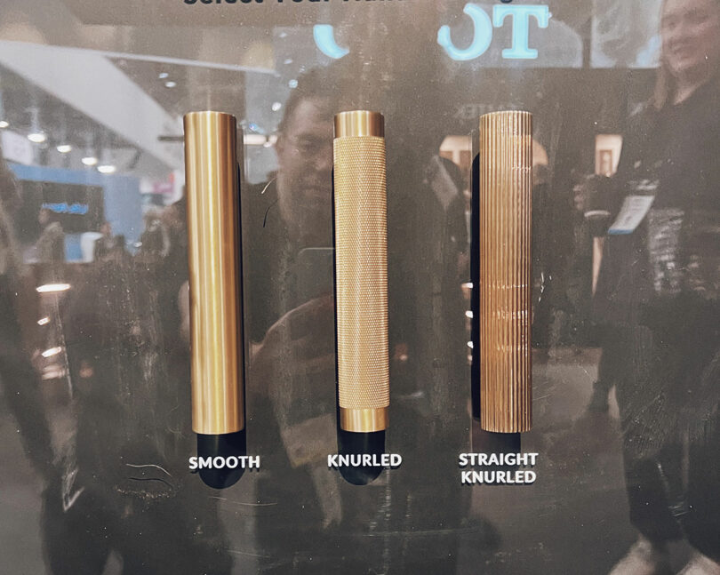 Three metallic bar grips displayed vertically: Smooth, Knurled, and Straight Knurled. Reflections and people visible in the background, reminiscent of a bustling KBIS exhibit.