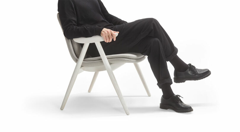 A person in black attire sits gracefully on a Keilhauer chair against a pristine white background, with legs crossed and hands resting elegantly on the armrests.