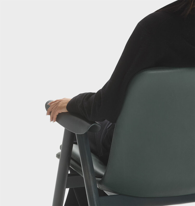 A person sits comfortably on a green Keilhauer chair, right hand gracefully resting on the armrest, wearing a sleek black top against a plain white background.