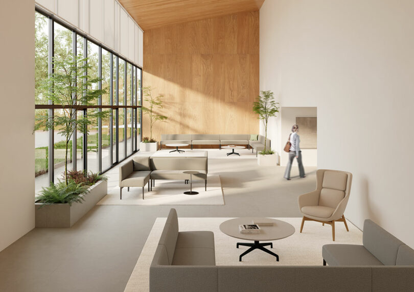 A modern, spacious lobby with large windows and Keilhauer's minimalist furniture showcases potted plants. A person strolls through the bright room, featuring neutral tones and a striking wood accent wall.