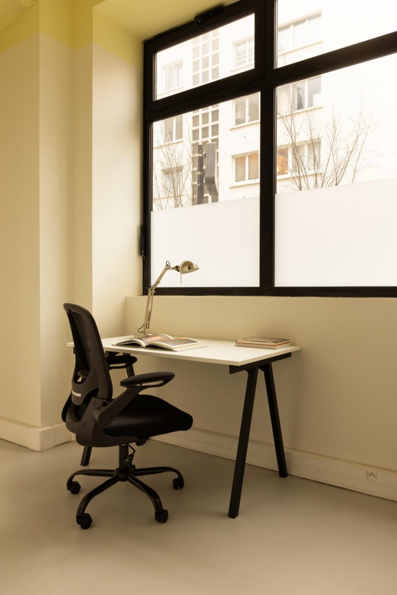 A minimalistic office setup with a black chair, a white desk, a desk lamp, and some papers in front of a large window.