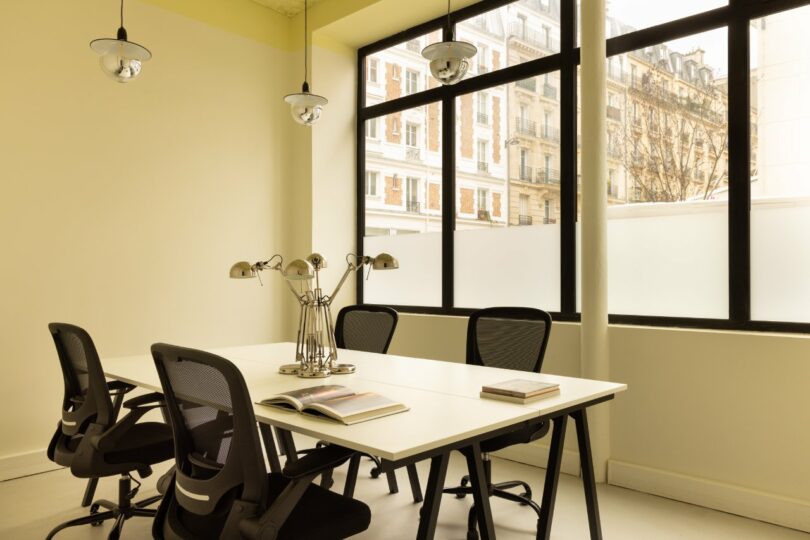 A modern conference room with four chairs around a rectangular table, featuring lamps and open books. Large windows reveal a view of an adjacent building.