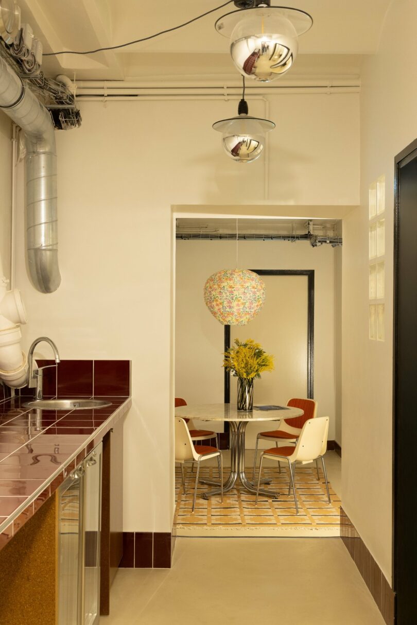 A corridor leads to a dining area. The foreground has a tiled counter and hanging lights, while the background features a round table, colorful pendant lamp, and chairs on a patterned rug.