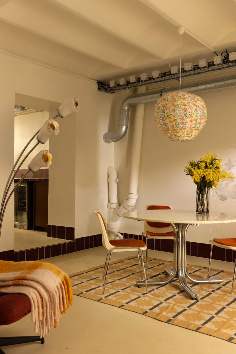 A modern dining area with a round table and chairs on a patterned rug. A floral pendant light hangs above, and a vase of yellow flowers adorns the table. A floor lamp stands nearby.