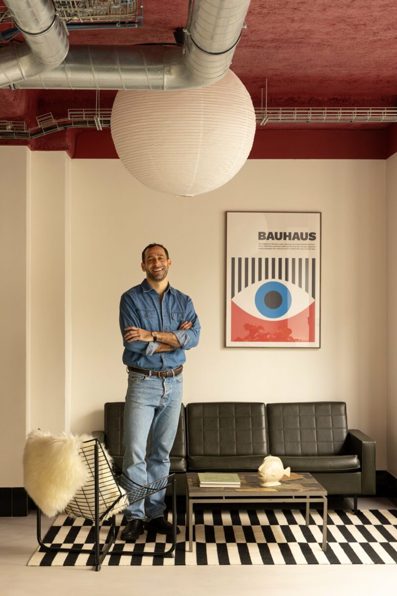 A person stands in a modern living room with a black sofa, patterned rug, and Bauhaus poster. They wear a denim shirt and jeans, standing next to a wire-frame chair with a fur cushion.