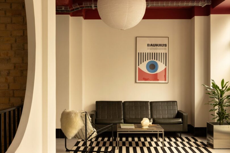 Stylish room with a black sofa, modern chair, striped rug, and a Bauhaus-themed artwork. Large round light fixture and a potted plant add to the decor.