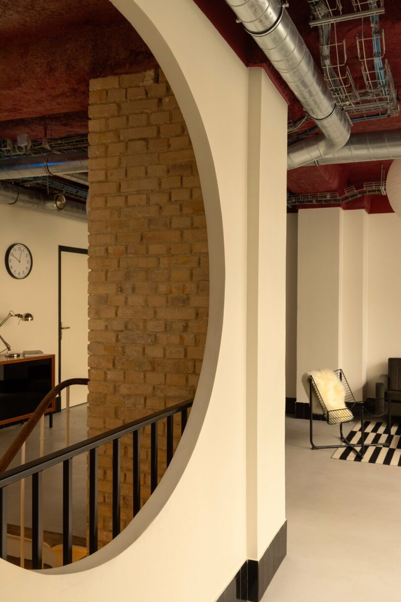 Interior with exposed brick column in circular cutout, industrial ceiling, clock, modern furniture, and black-and-white rug.