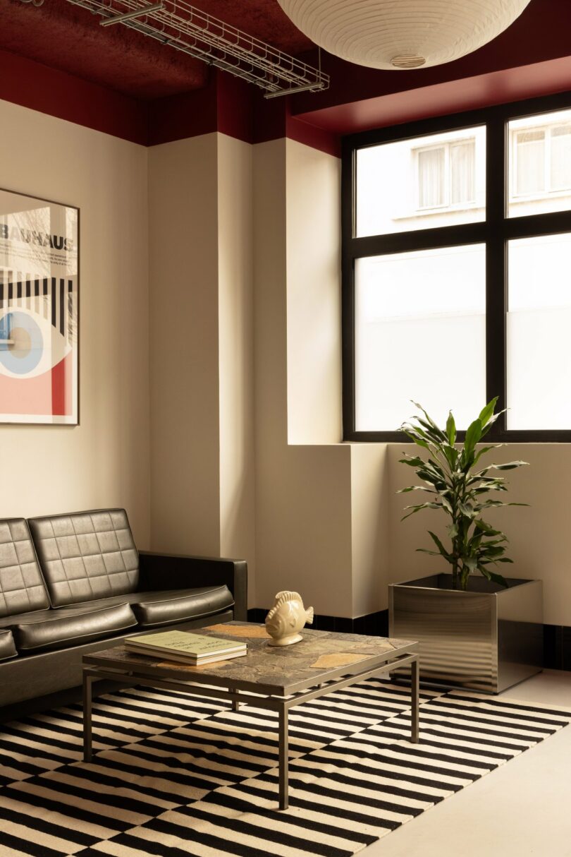 Modern living room with a black leather sofa, striped rug, coffee table, potted plant, and large window. Abstract wall art, white walls, and red ceiling add contrast to the space.