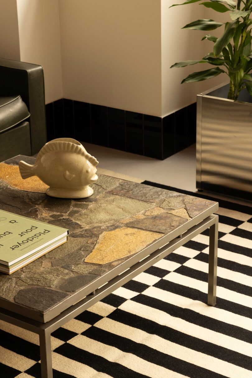 A living room with a stone-top coffee table, fish-shaped sculpture, striped rug, black leather chair, and a tall plant in a metal planter.