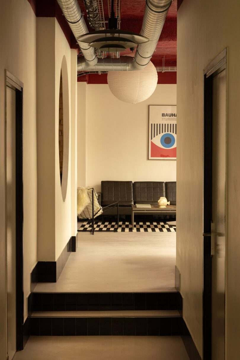 Hallway with two doors leading to a room. The room features black sofas, a checkered floor, and a round hanging light. A geometric art piece is mounted on the back wall.