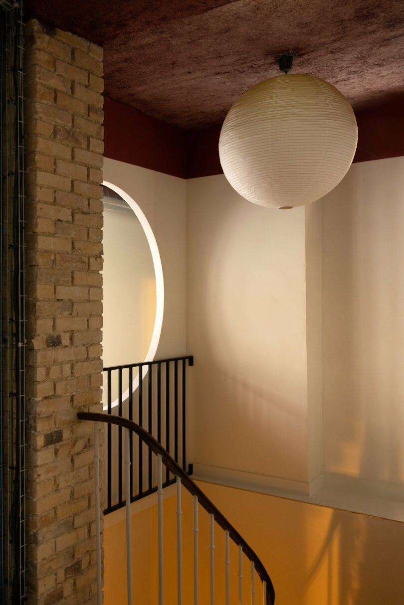 A stairway with a curved handrail descends beside a brick wall. A round paper lantern hangs from the ceiling, above a railing and a circle-shaped wall feature.