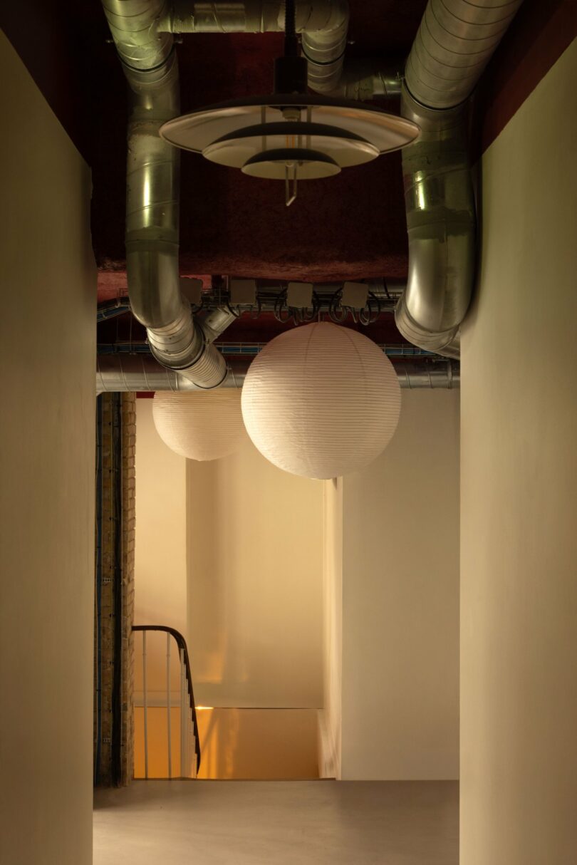 A narrow hallway with exposed industrial pipes on the ceiling and round paper lanterns. A staircase with a metal handrail is visible in the background.