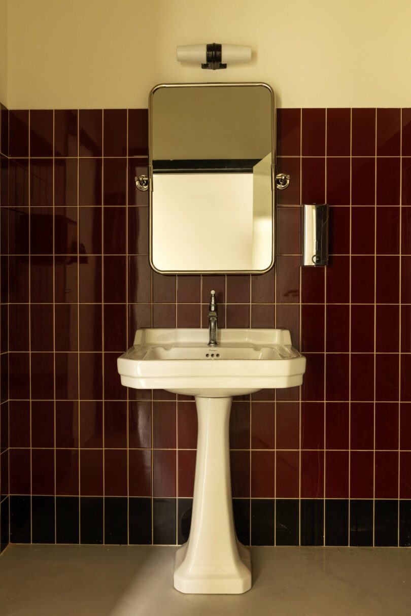 A white pedestal sink with a mirror above it is mounted on a wall covered with dark red tiles. A small light is positioned above the mirror.