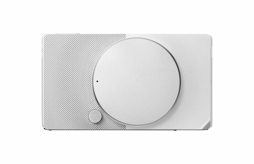 The Sigma BF is a minimalist white rectangular device featuring a large circular dial and a smaller button. Its surface boasts a textured design on the left side, adding an elegant touch to its sleek appearance.