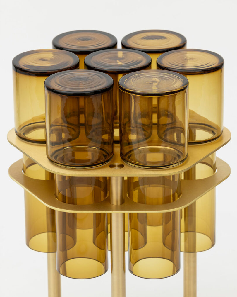 A cluster of eight cylindrical amber glass containers, joined in two layers, is supported by a brass-colored metal frame.