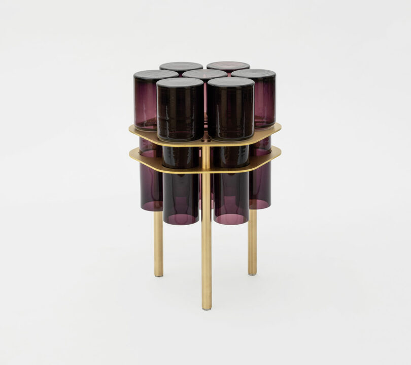 A sculpture joins two tiers of cylindrical purple glass containers, nestled within a gold framework stand, creating a stunning visual field.