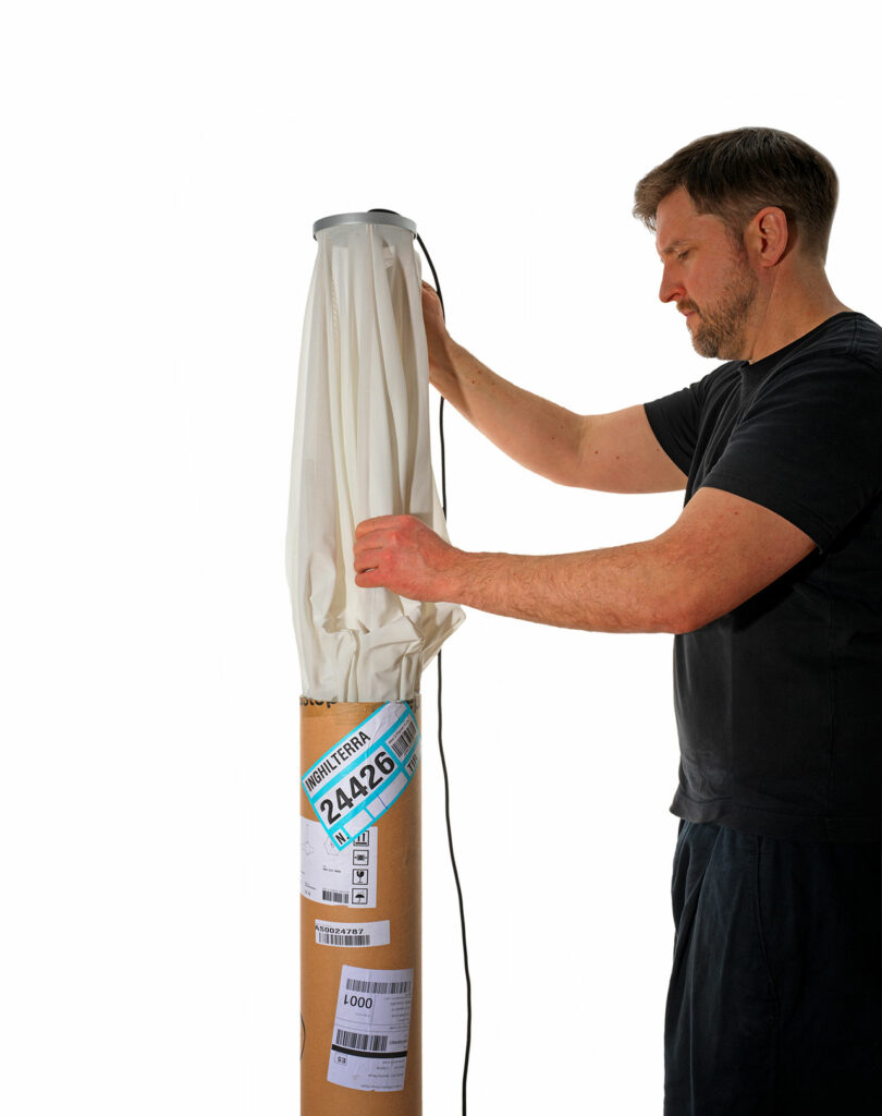 A man carefully arranges a light diffuser on a tall cylindrical stand, the label showing shipping details. Designed by Samuel Wilkinson, this piece is sure to add an element of sorpresa to any setting with its innovative flair.