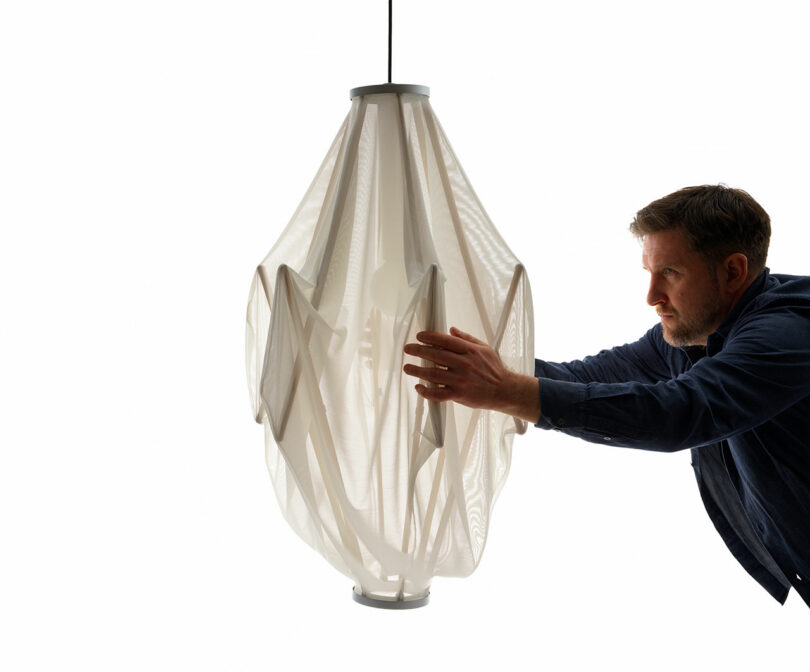 A man adjusts the large Sorpresa pendant light fixture, a Samuel Wilkinson design, featuring a draped, translucent fabric aesthetic set against a pristine white background.