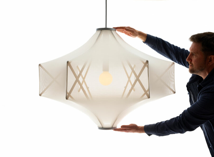 A man adjusts the striking Sorpresa pendant lampshade, designed by Samuel Wilkinson, featuring a large white geometric form with a visible light bulb, set against a pristine white background.