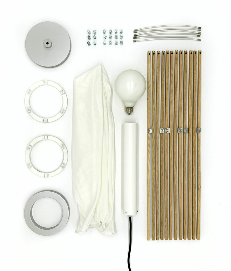 The Sorpresa lamp assembly kit, designed by Samuel Wilkinson, includes wooden rods, a light bulb, fabric shade, metal frame rings, ceiling mount, screws, and a power cord—all meticulously arranged on a white background.