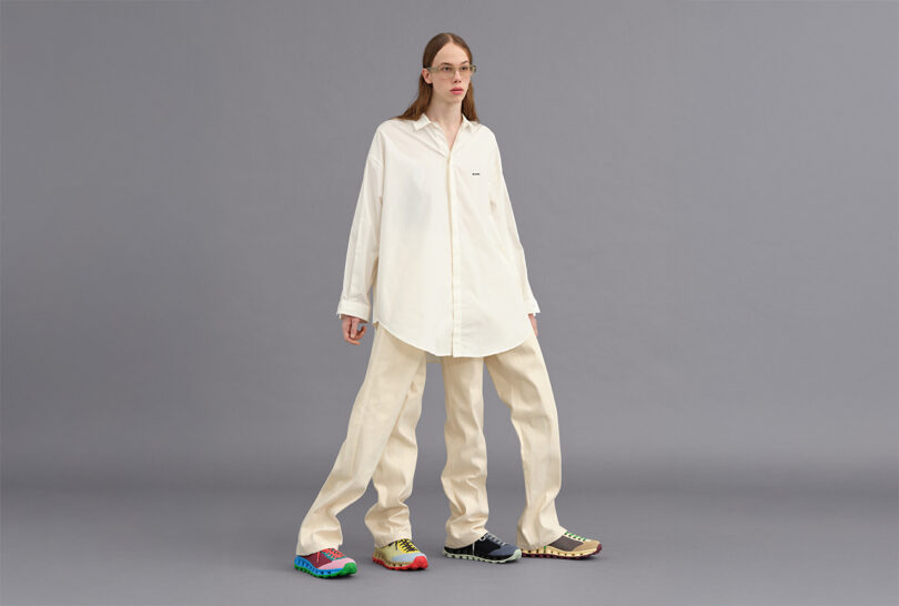 Person wearing oversized white shirt and beige trousers, standing against a plain gray background. They have colorful shoes and are looking ahead.