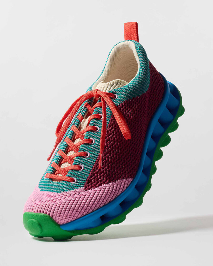 Colorful sneaker with a red mesh upper, turquoise lace eyelets, orange laces, and a blue and green wavy sole.