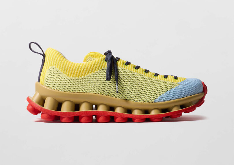 Yellow and blue knit sneaker with black laces, red cushioned sole, and brown midsole on a gray background.