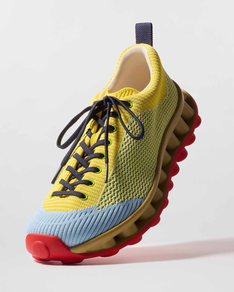 Multi-colored sneaker with a mesh upper in yellow, green, and blue, black laces, and a red sole with a unique, wavy tread design, displayed against a plain background.