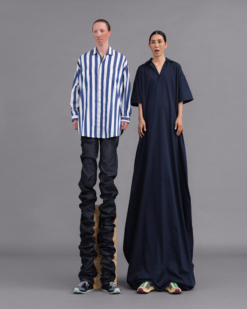 Two individuals stand against a gray background wearing exaggerated, oversized clothing. One wears a striped shirt and long jeans; the other wears a long navy dress. Both wear sneakers.