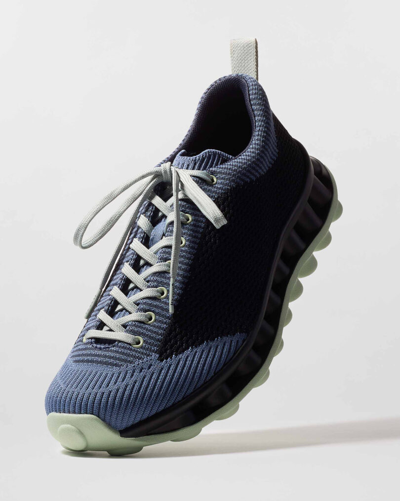 Close-up of a modern athletic shoe with a textured black and blue upper, gray laces, and a light green sole featuring a unique, rounded design.