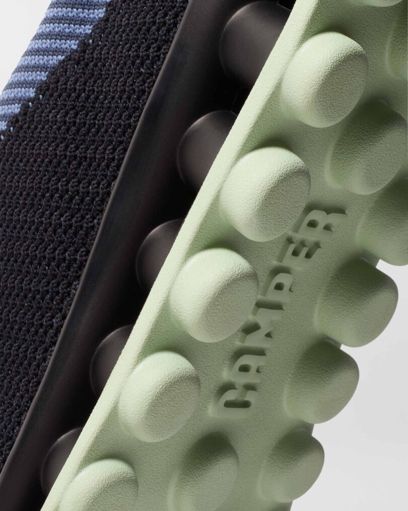 Close-up of a black sneaker with a textured green sole featuring round protrusions and the word "Camper" embossed on it.