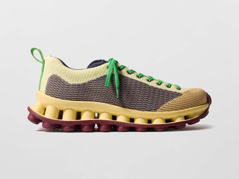 A multicolored sneaker with a knitted upper, green laces, and a thick, maroon and yellow wavy sole on a plain background.