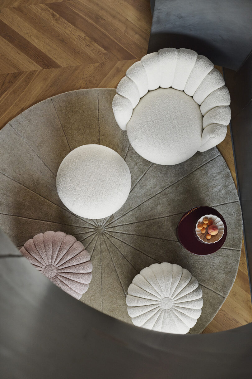 Top view of a room with a round beige rug, white plush chairs, a purple cushion, and a dark round table with a bowl of fruit on a wooden herringbone floor.